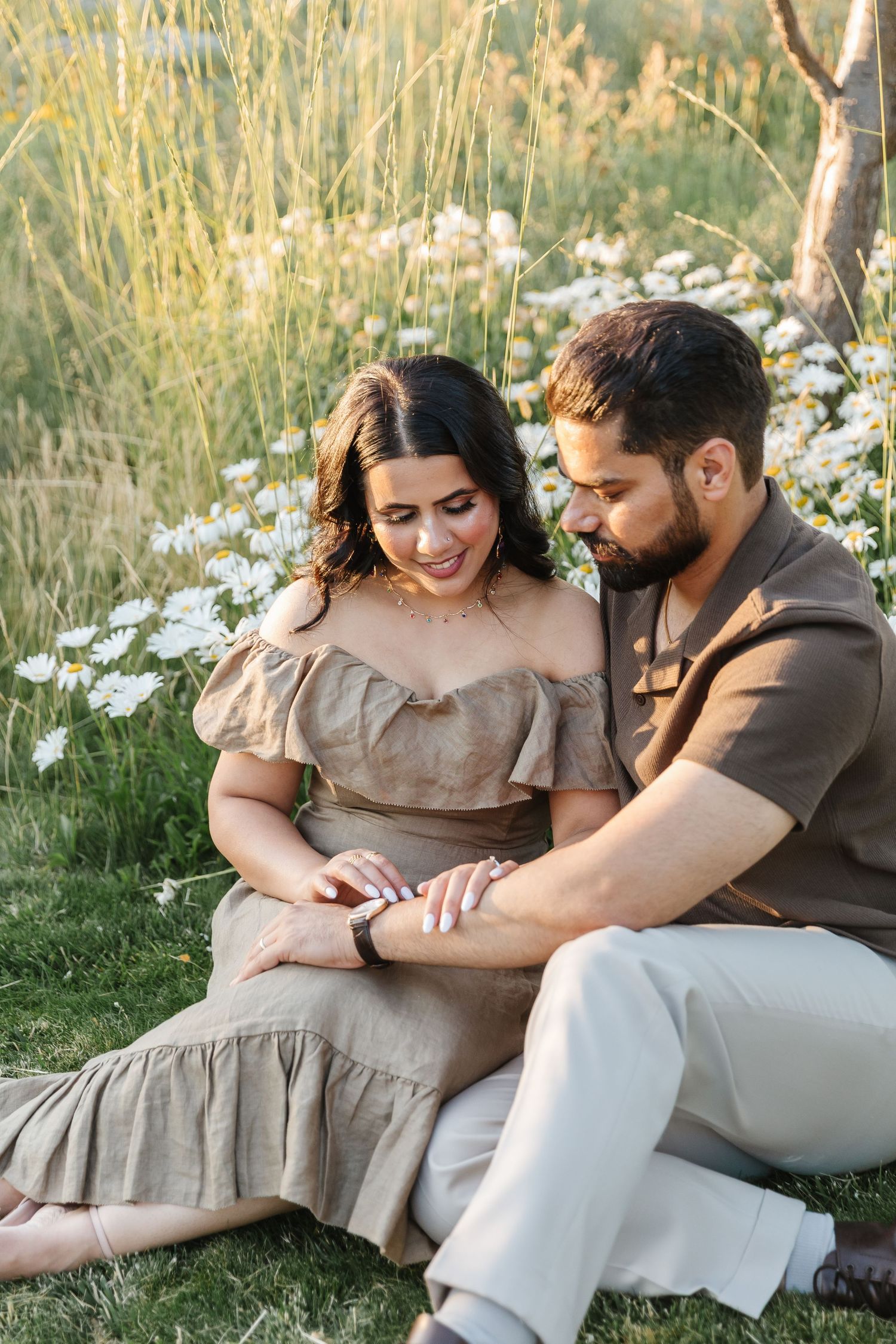 A couple sits together on grass with white flowers in a romantic outdoor setting at Sanctuary Gardens in West Kelowna for engagement photo's