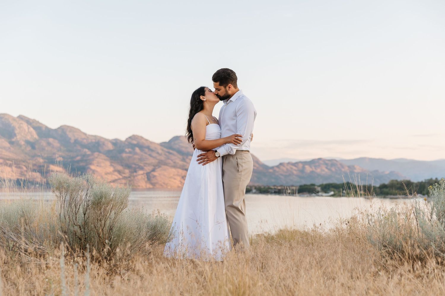 A couple in wedding attire take engagement photo's in a golden field at sunset with mountains in the background in West Kelowna, BC.