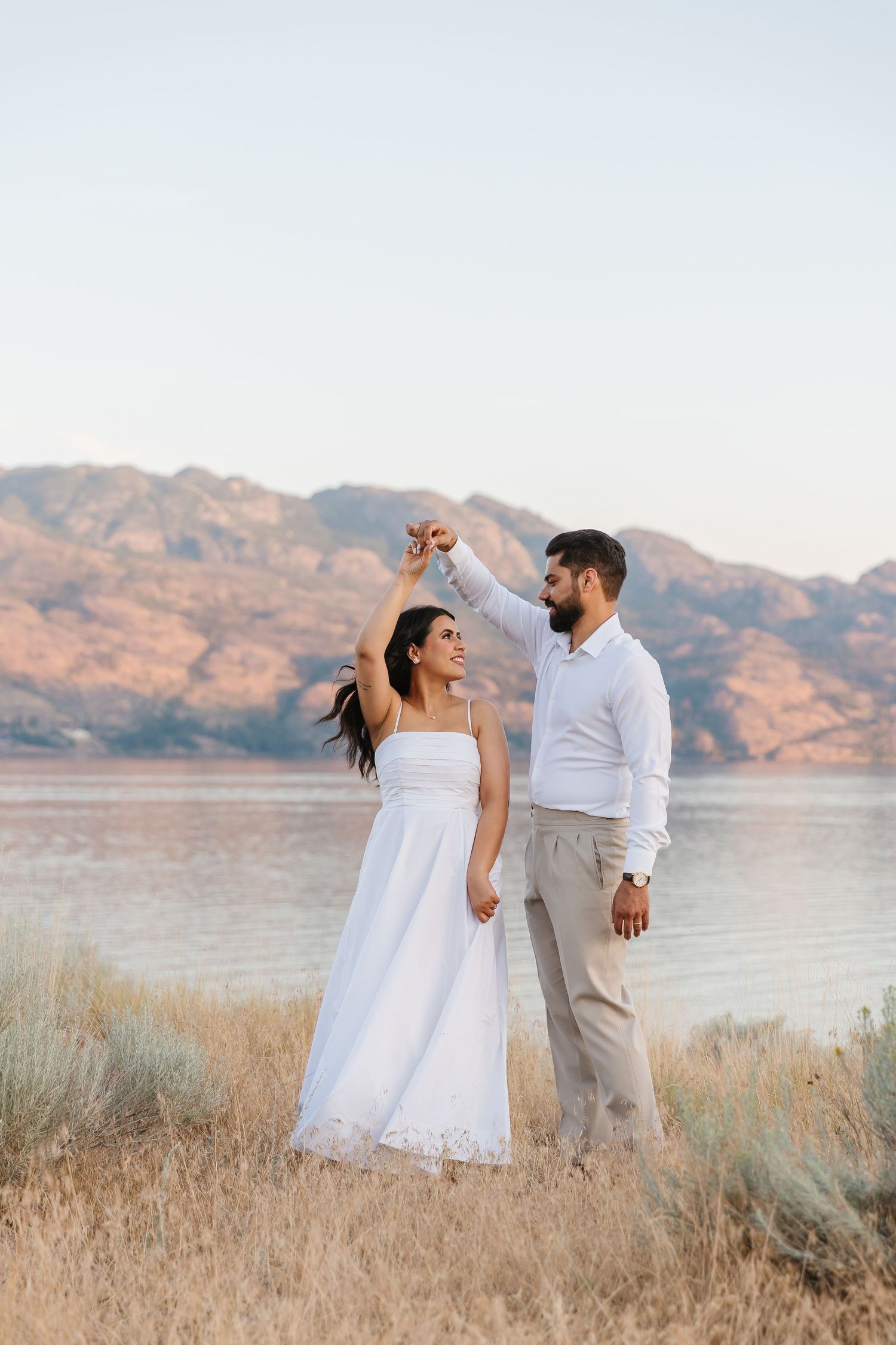 A romantic couple in white and beige dance together in a field in West Kelowna with mountains and Okanagan Lake in the background