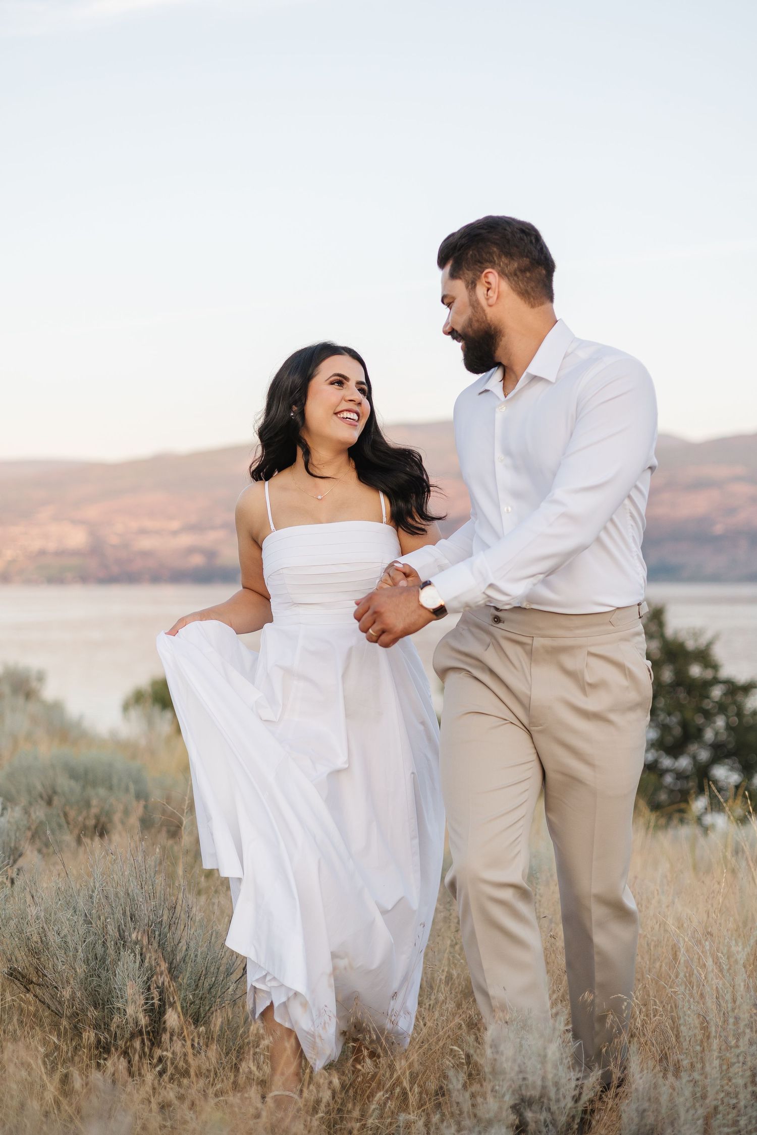 A couple in matching white outfits dance playfully in a golden field at Sanctuary Gardens in West Kelowna, BC with mountain views.
