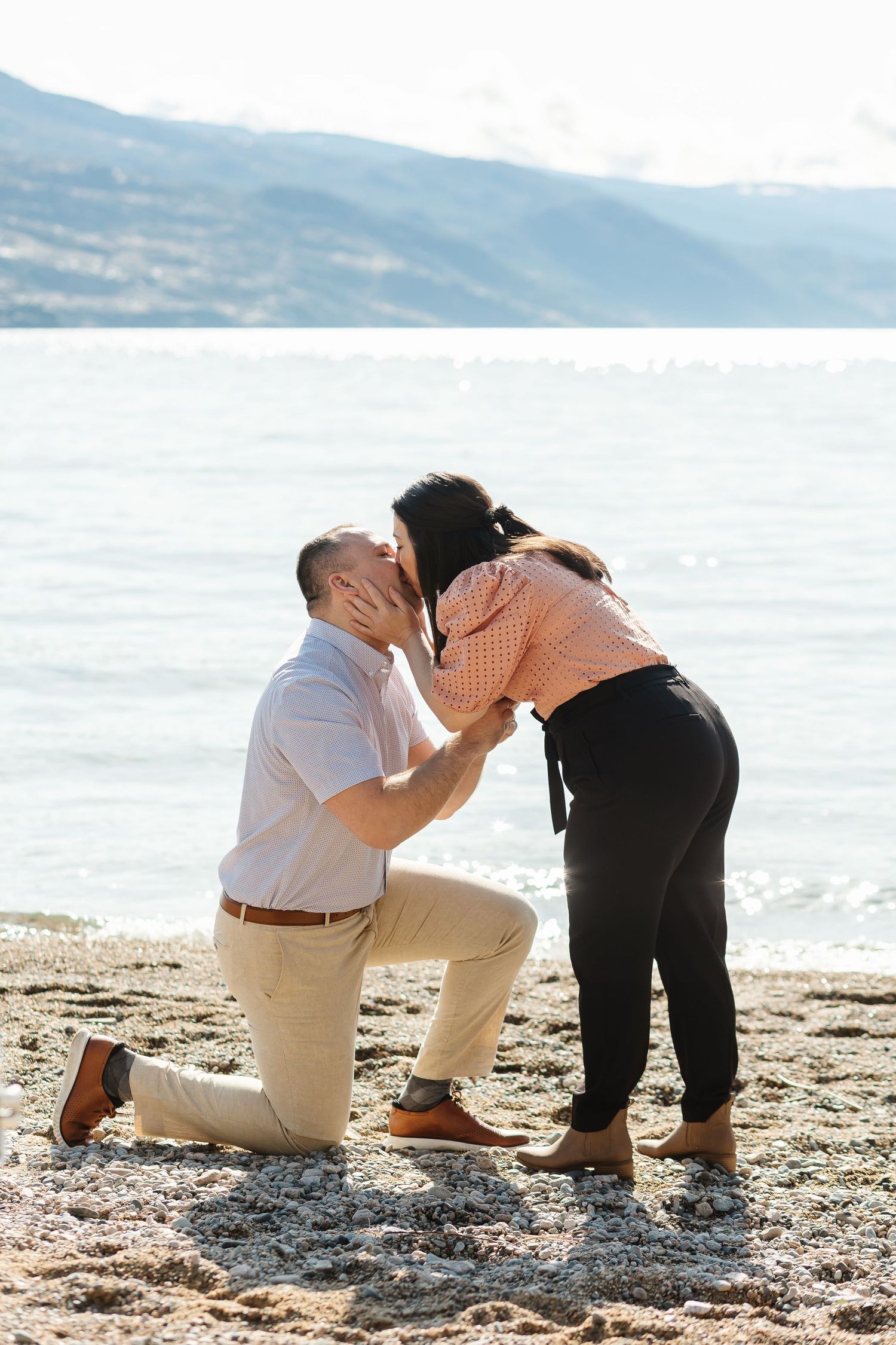 Couple hugging on the beach in Kelowna BC during an engagement session with Okanagan lake and the mountains in the background.