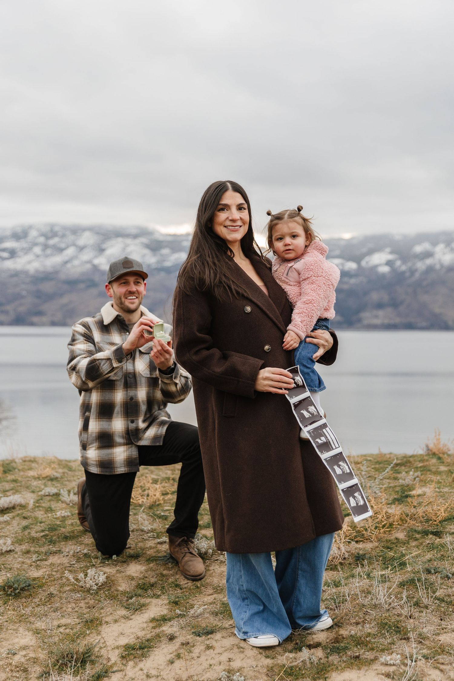 A family poses together on a hillside overlooking a scenic Okanagan Lake in the background during a surprise proposal photoshoot in Kelowna.