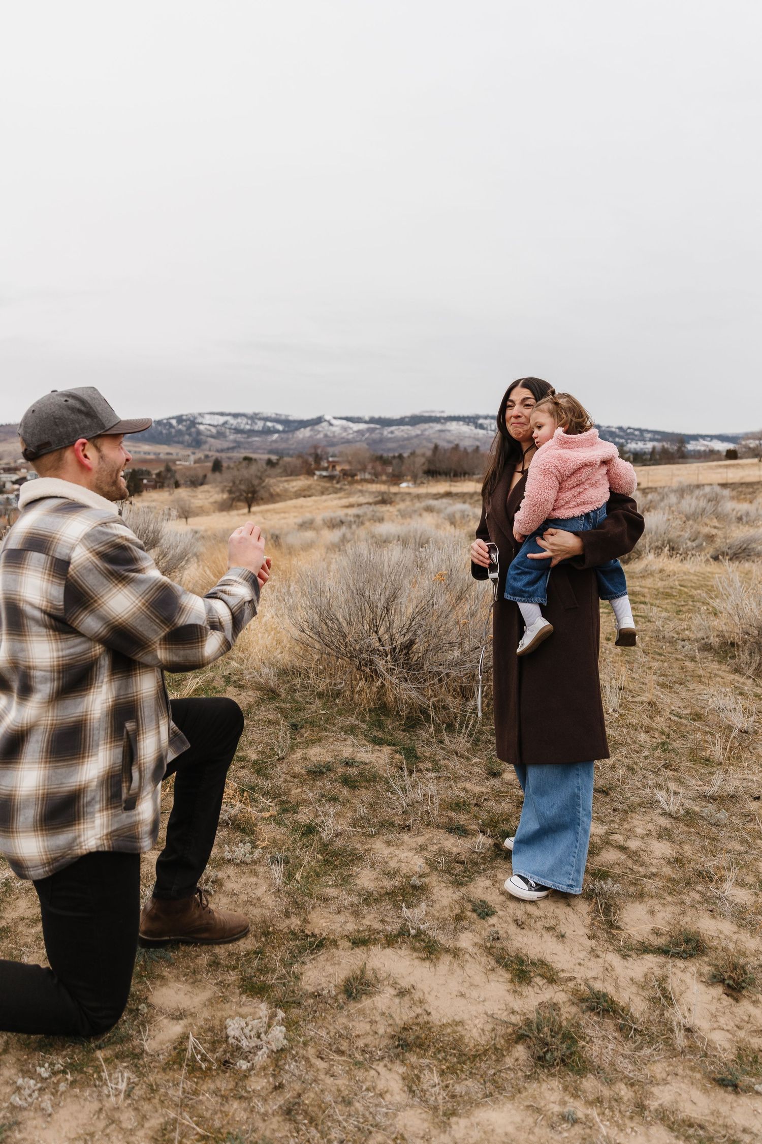 A couple shares a moment in a field with brown grass hills in the background during surprise proposal photoshoot in West Kelowna, BC.