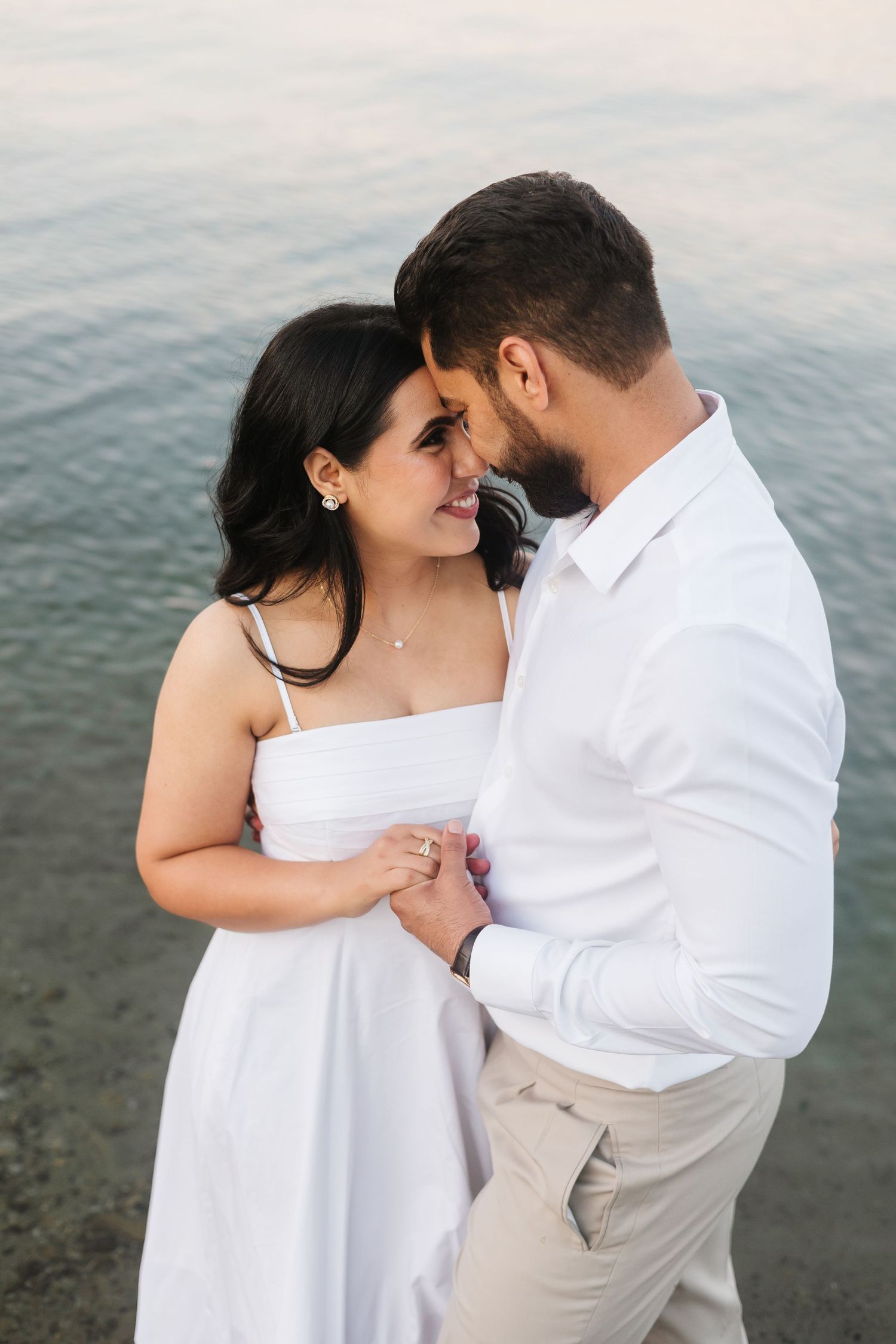 A romantic couple shares an intimate moment on a beach wearing white and beige outfits in West Kelowna, BC for an engagement photo session.