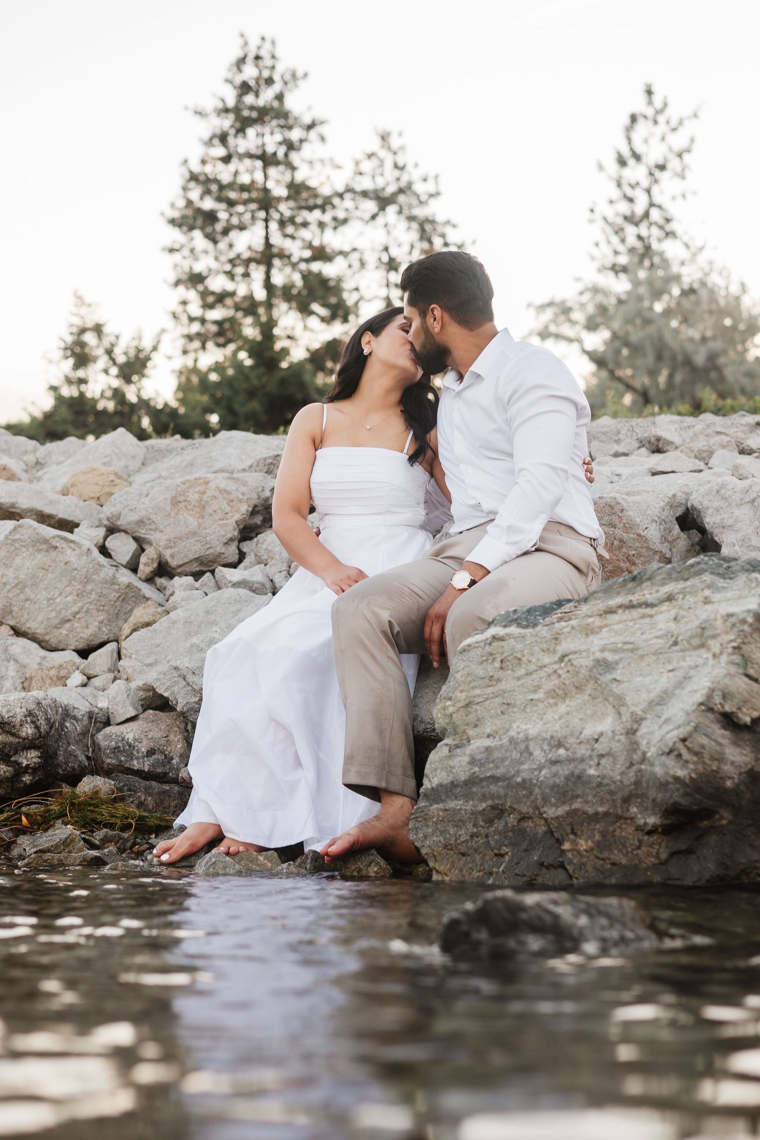 A couple shares an intimate moment while sitting on rocky shoreline by the water in Okanagan Lake for engagement photos.