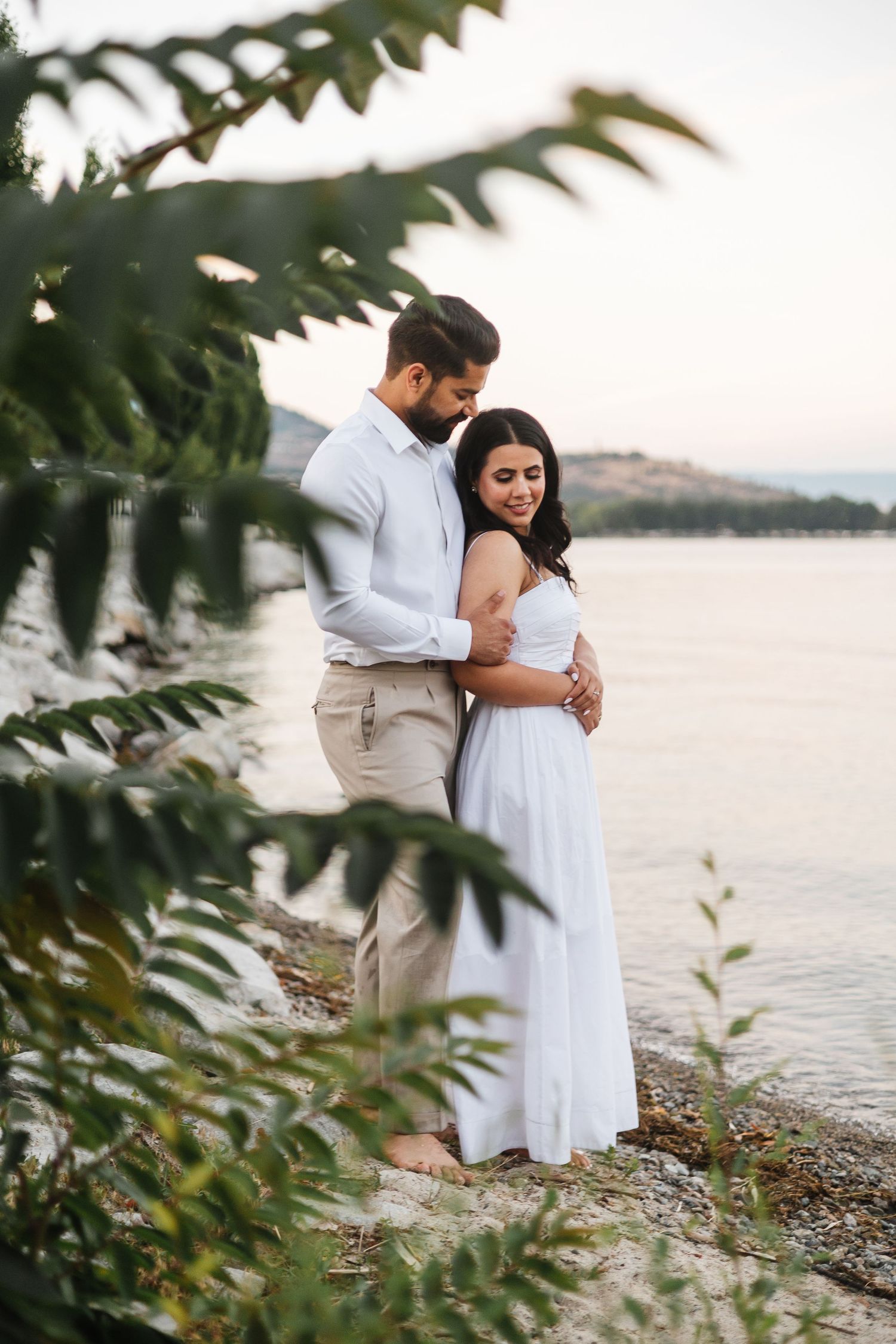 Engagement photos on the beach in West Kelowna with greenery and a view of Okanagan Lake and the mountains at sunset