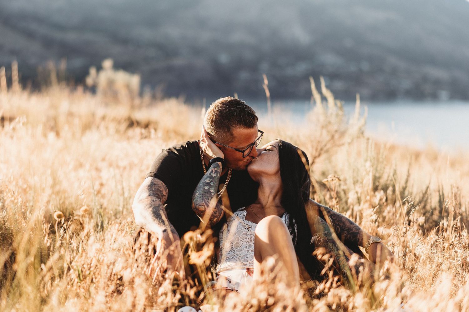 Couple sitting in golden wheat field during sunset with mountains in background at Knox mountain for an engagement photoshoot