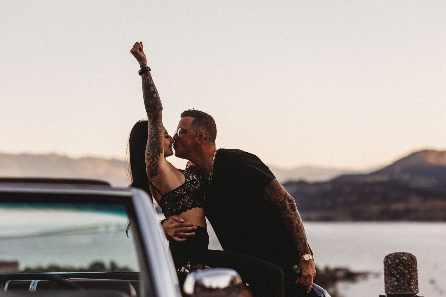 Romantic sunset scene with couple sitting on a vintage car overlooking a mountain lake view celebrating their engagement in the Okanagan.