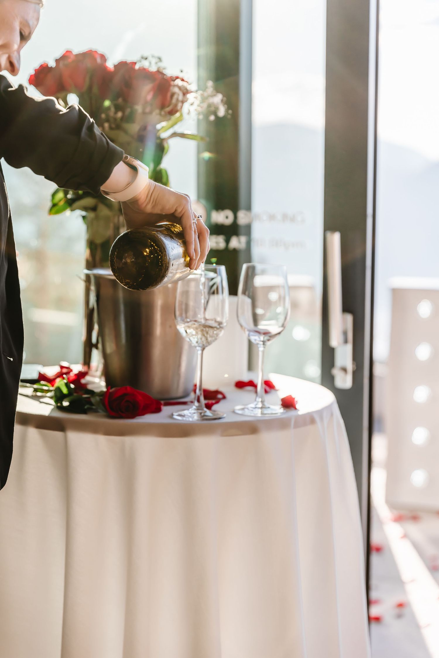 Someone pours champagne into wine glasses on an elegant white tablecloth decorated with red rose petals after surprise proposal engagement
