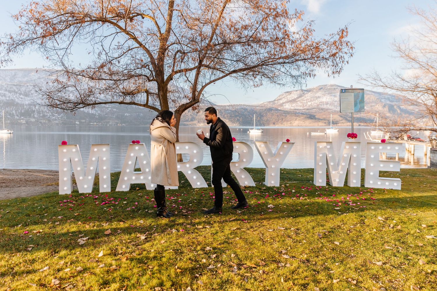 A romantic surprise proposal photography session under trees with large white marquee signs letters spelling out MARRY ME in Kelowna BC