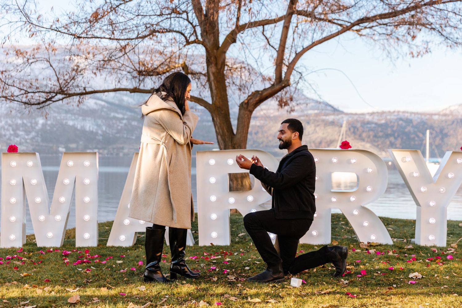 A romantic surprise proposal photography session under trees with large white marquee signs letters spelling out MARRY ME in Kelowna BC