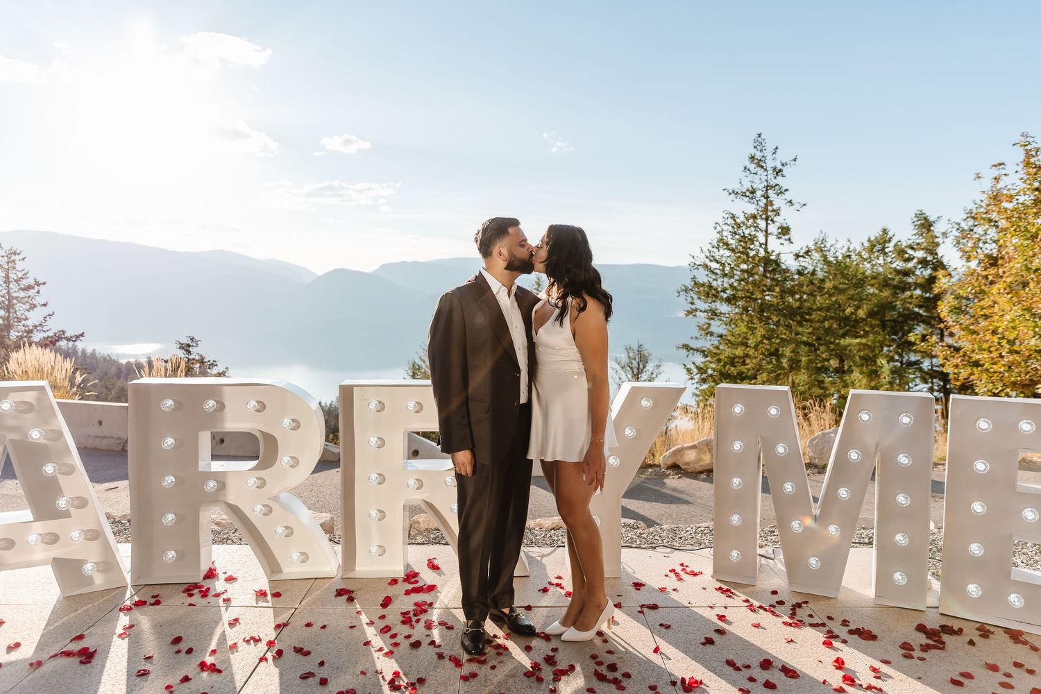 A couple shares a romantic kiss on an outdoor terrace with large white marquee letters and mountain views in the background in kelowna bc