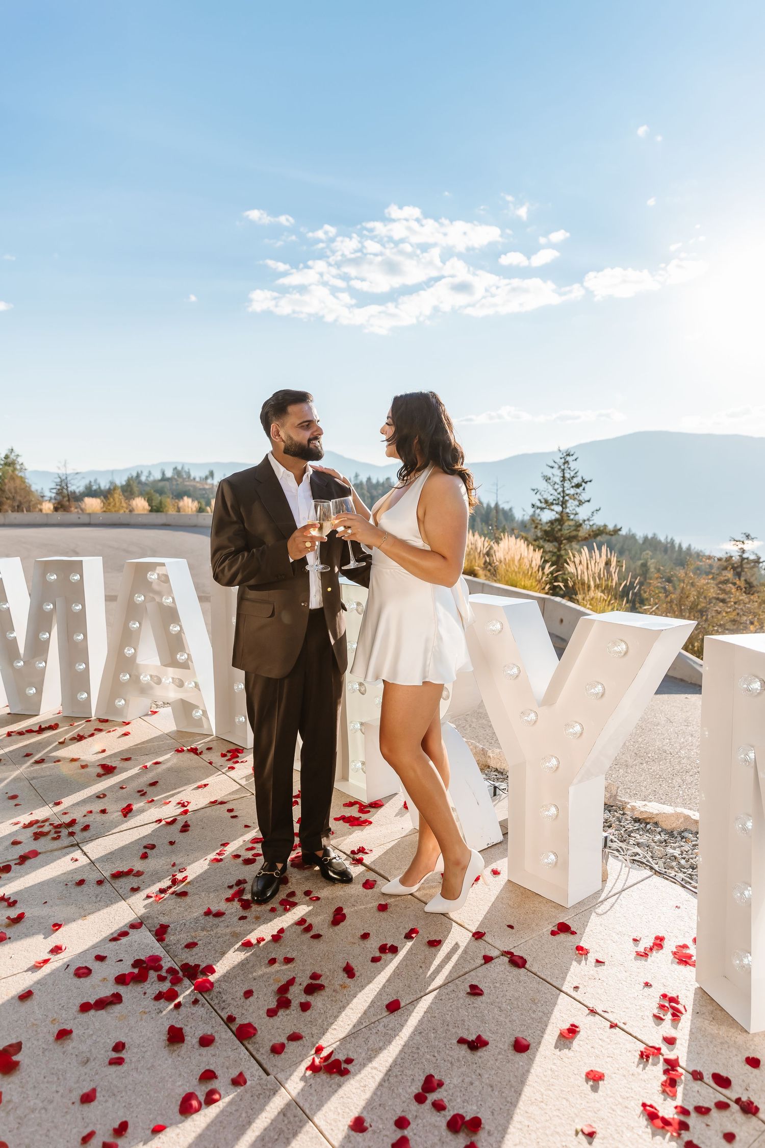 A couple celebrates with champagne in the Okanagan with view of mountains and marry me marquee letters after surprise proposal in kelowna bc