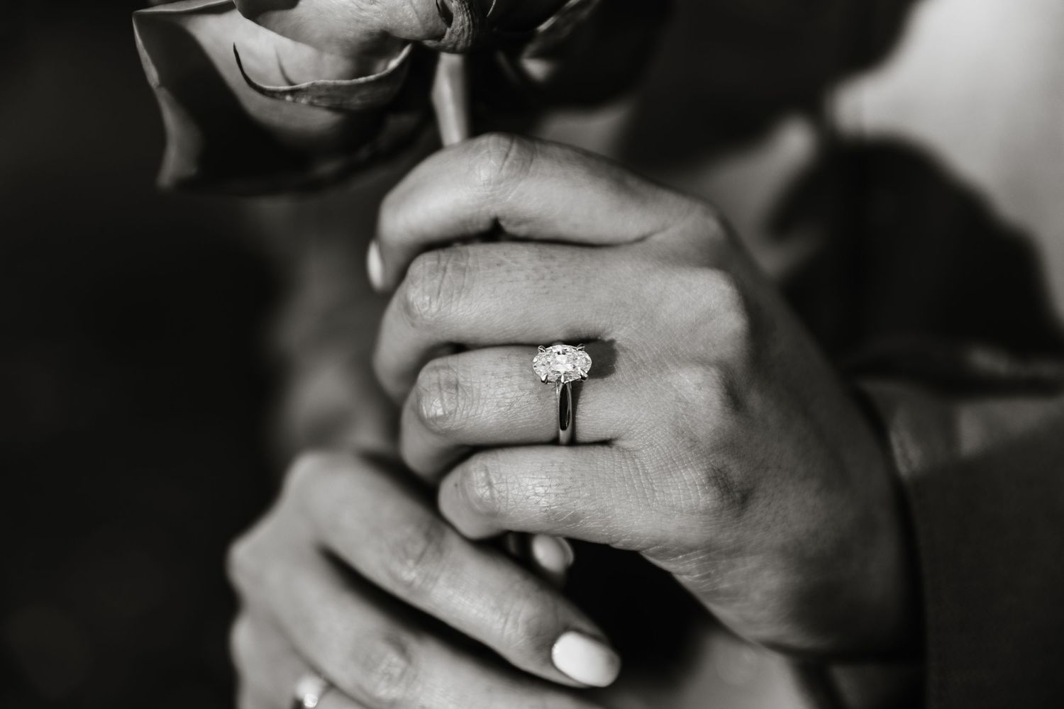 A close-up black and white photograph shows hands clasped together with a diamond engagement ring in Kelowna bc for a proposal session