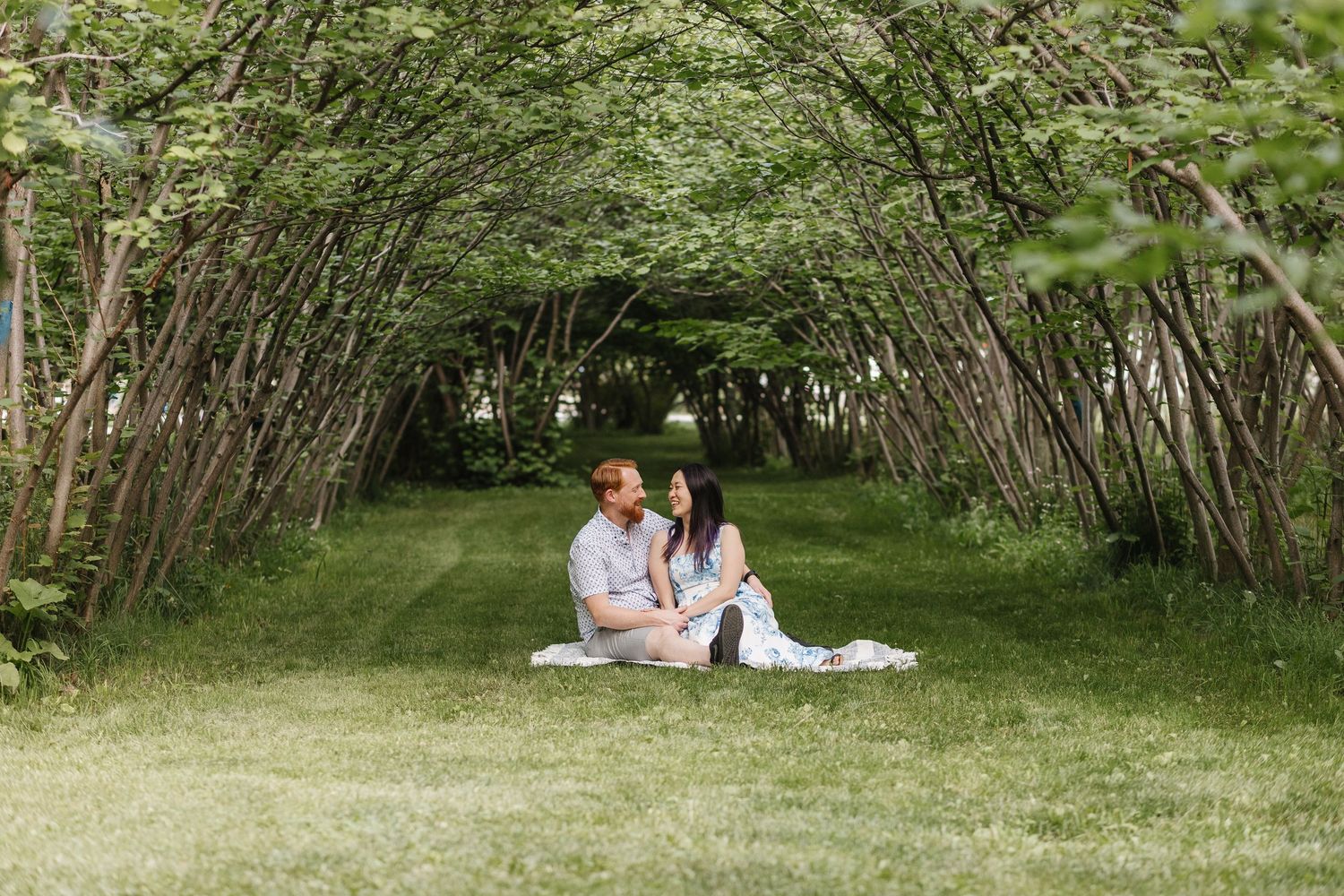 A romantic couple's photos sitting under archway of green trees in a serene outdoor setting in West Kelowna at Gellatly Nut Farm