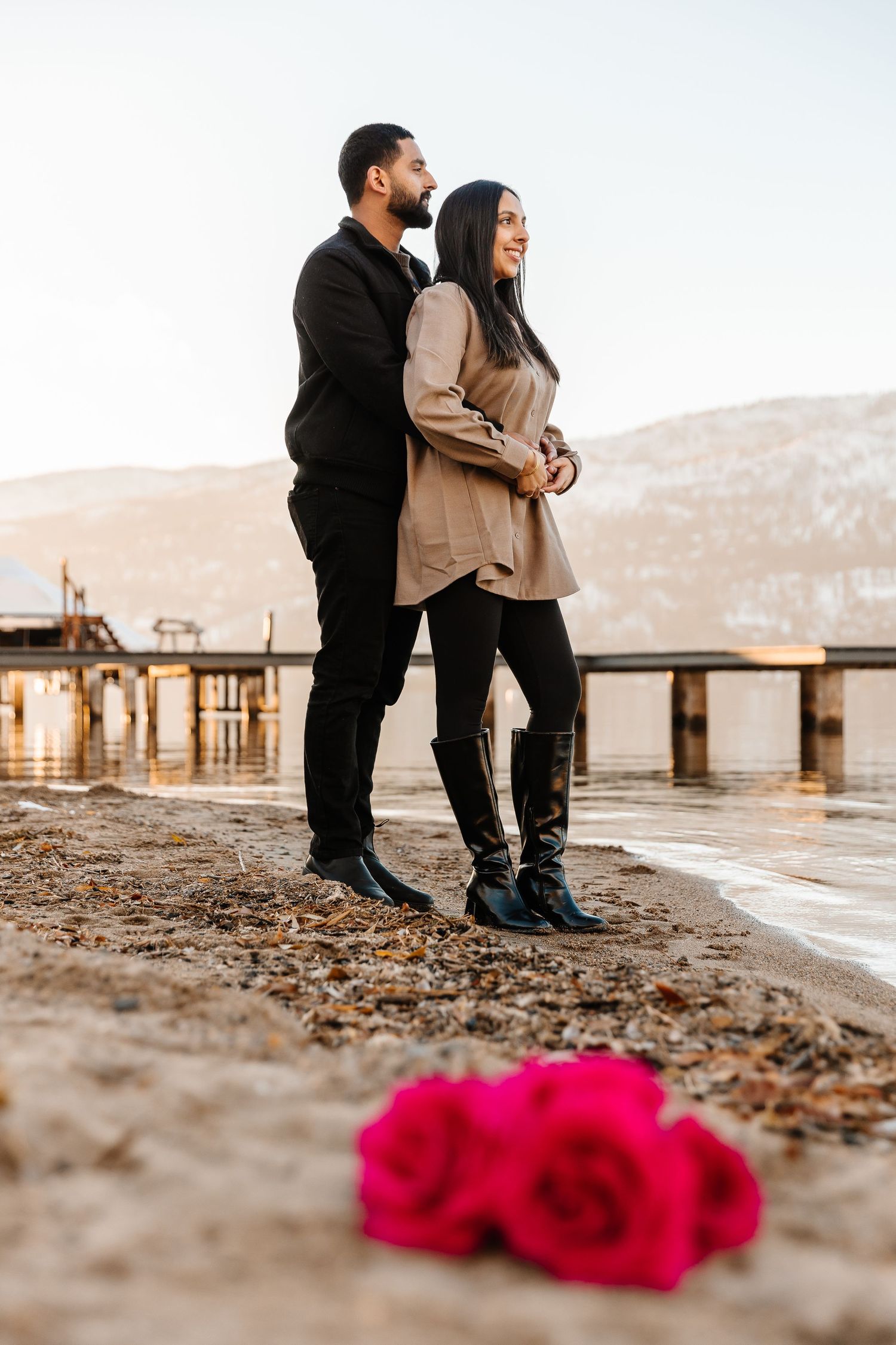 A romantic couple embraces on a lakeside beach with red roses in the foreground and Okanagan Lake for a surprise proposal in Kelowna BC