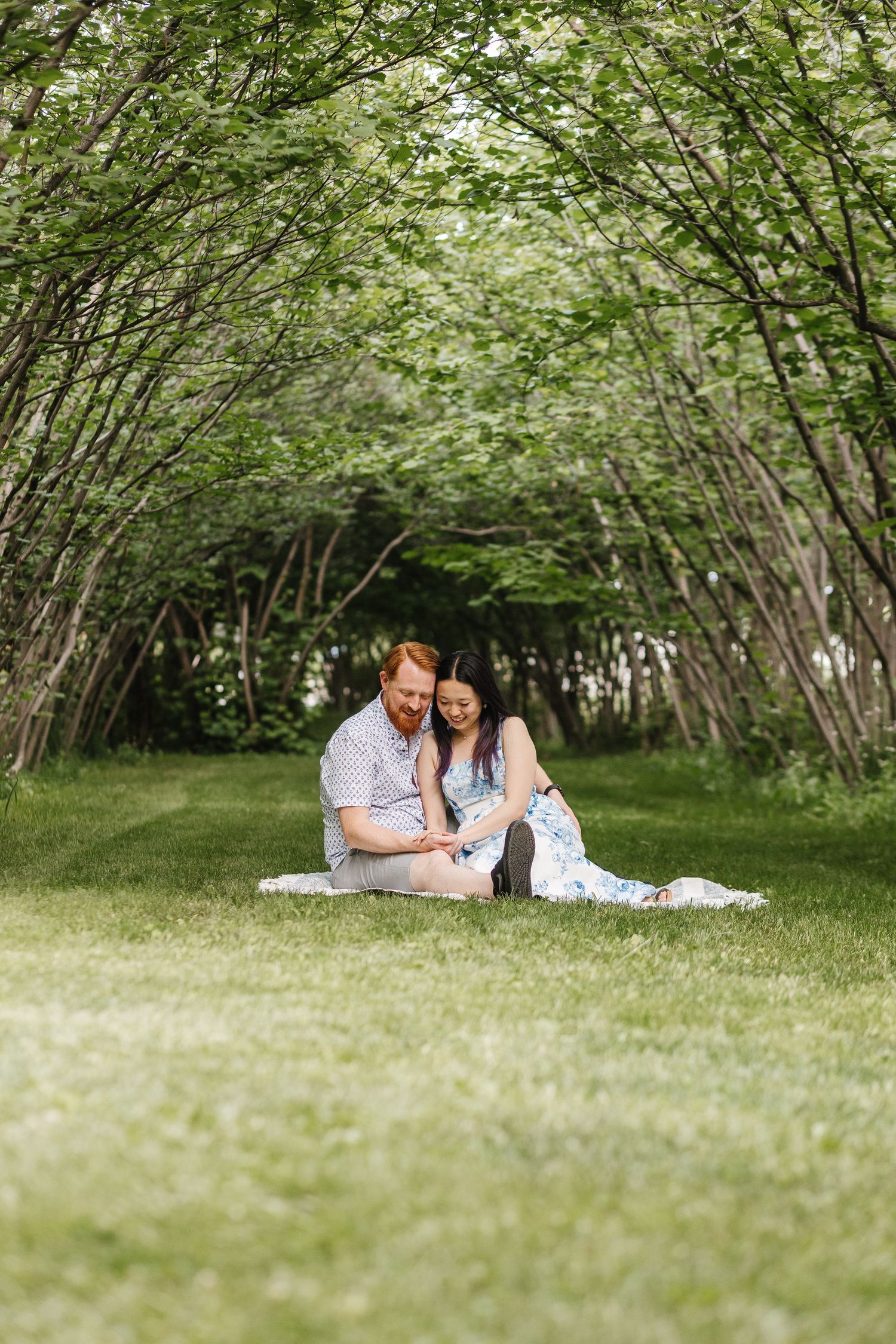 A romantic couple's photos sitting under archway of green trees in a serene outdoor setting in West Kelowna at Gellatly Nut Farm