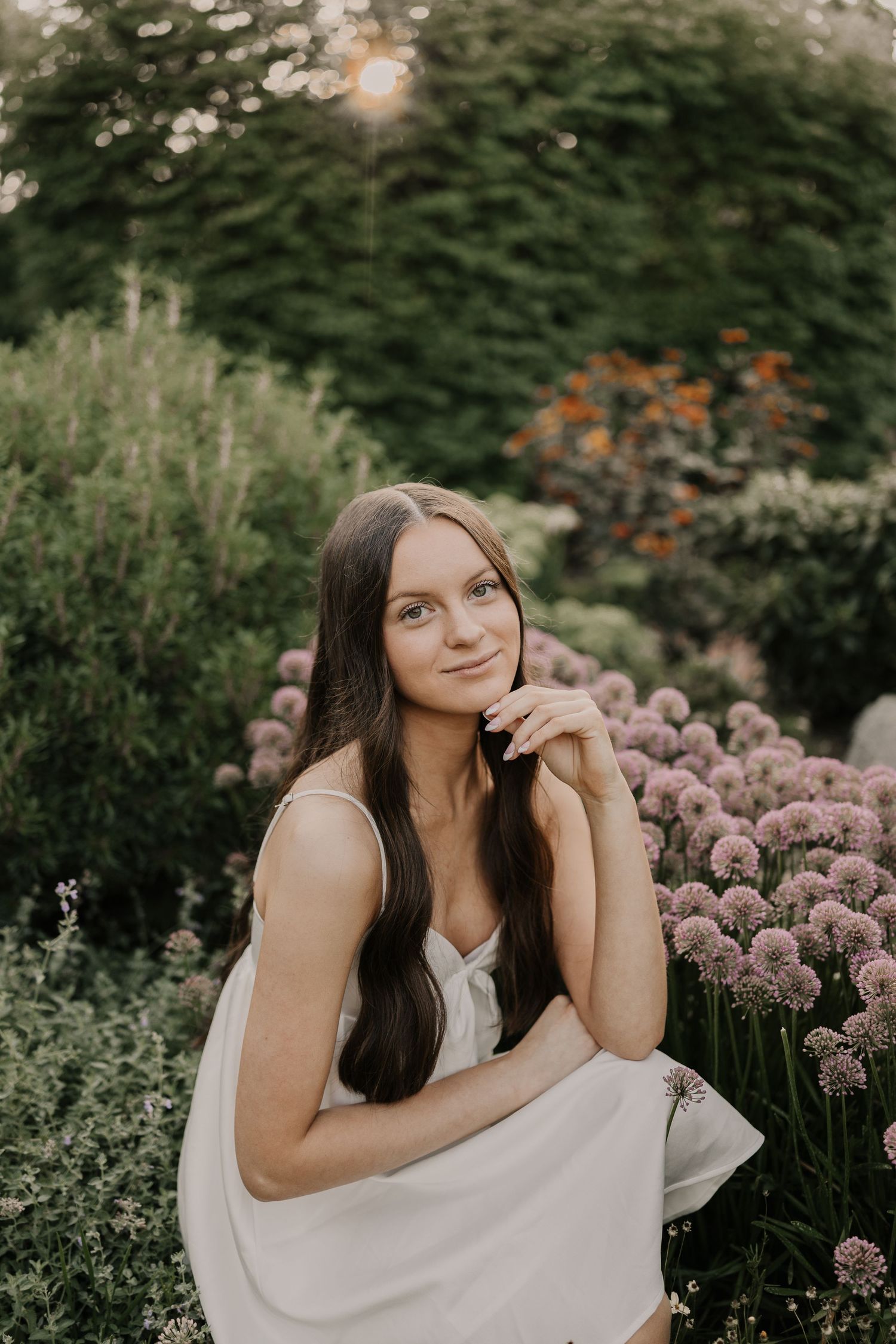A woman in a white dress poses gracefully among blooming purple allium flowers in a garden setting.