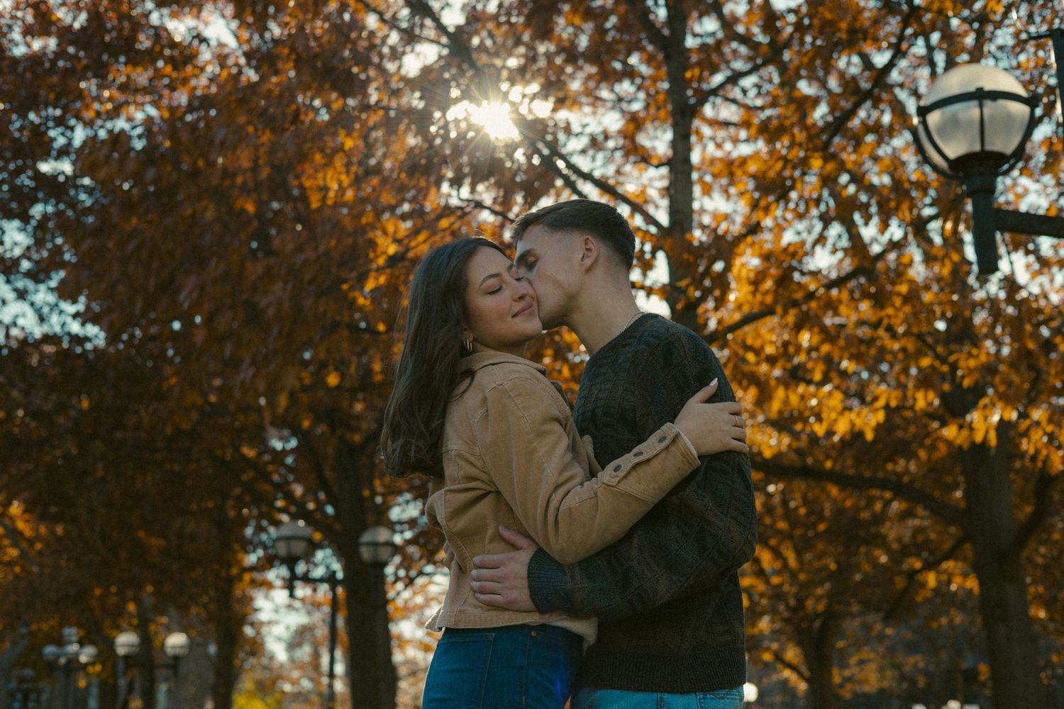 A romantic couple embraces during golden hour beneath autumn trees during a park engagement session in Ann Arbor Michigan.