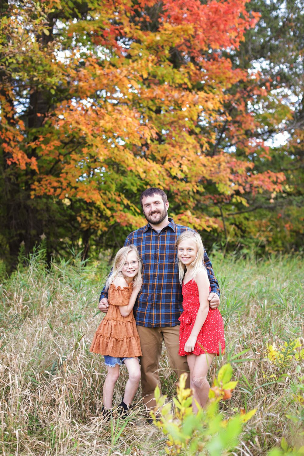 A couple poses together in a field during autumn with vibrant orange and red fall foliage in the background.