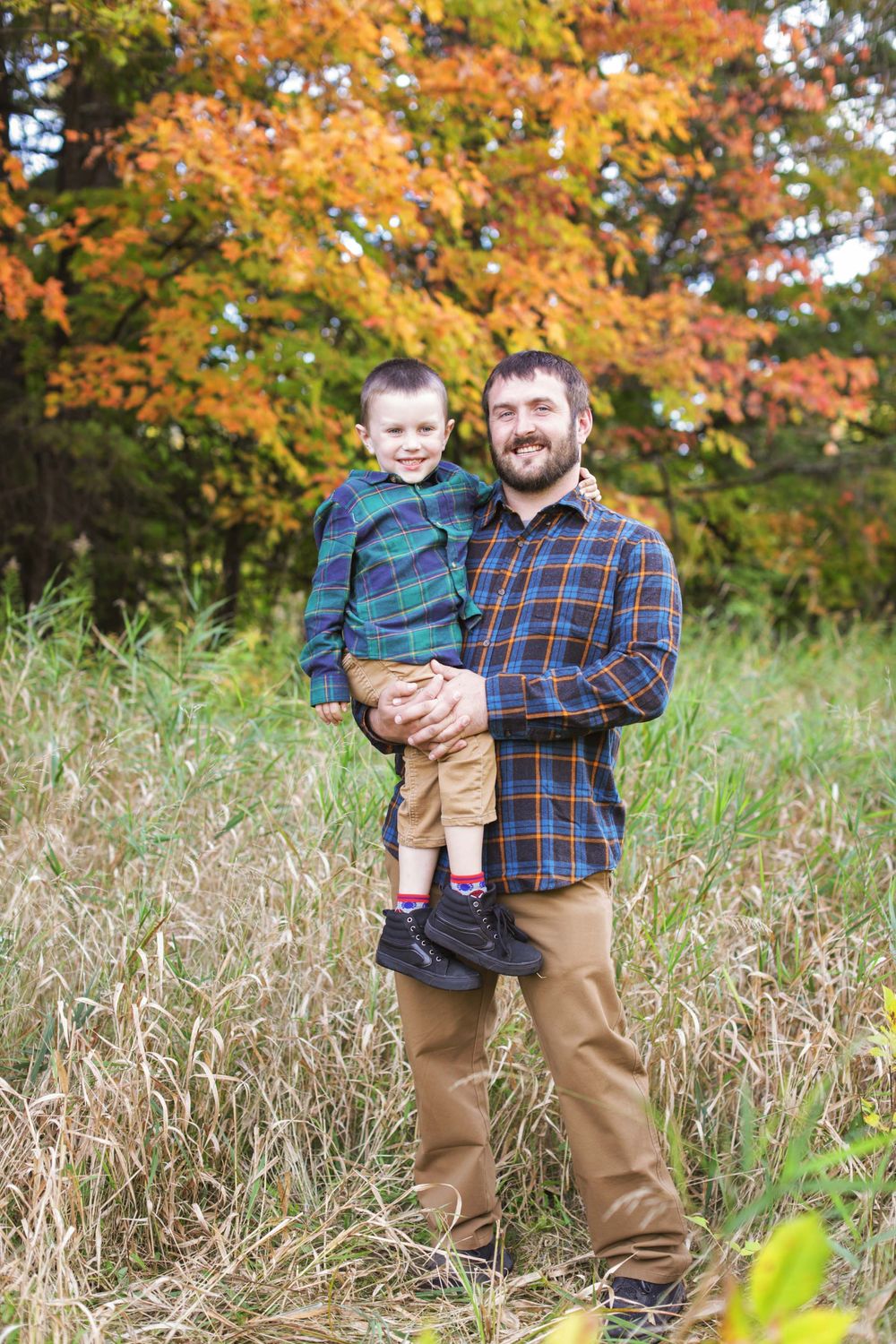 Adult and child stand together in a field of tall grass with vibrant autumn foliage in the background.
