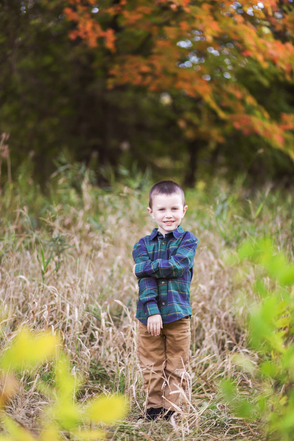 A child in a plaid flannel shirt and khaki pants stands in a field of tall autumn grass with orange foliage in background.