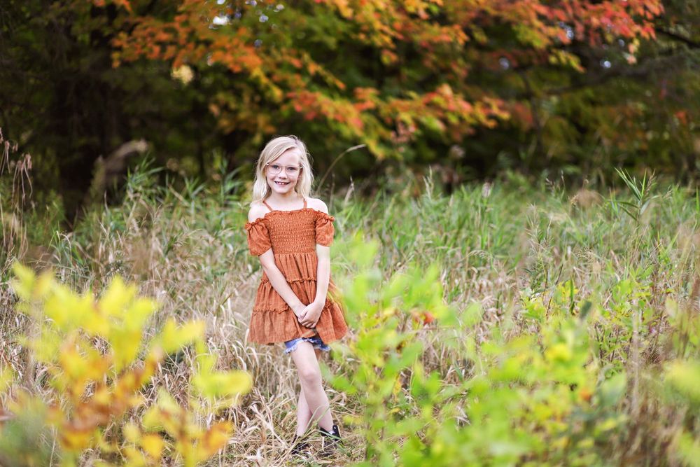 A person in an orange top stands in a golden autumn meadow with wildflowers and tall grass at sunset.