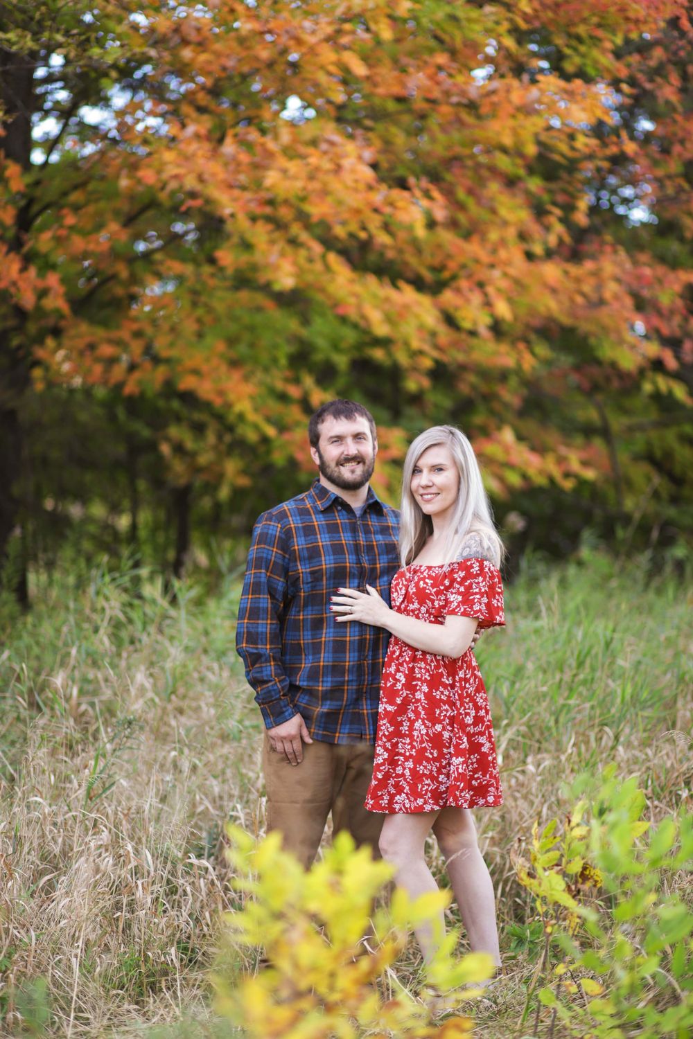 A couple stands close together in a grassy meadow with colorful autumn trees creating a scenic backdrop.