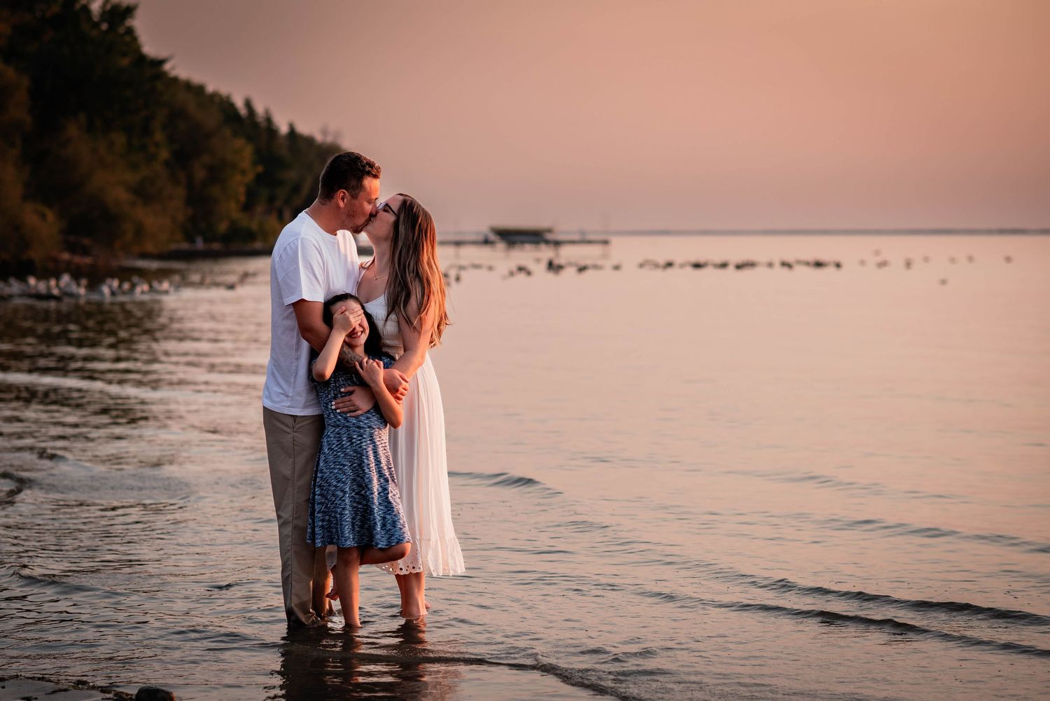 A recently engaged couple kissing at sunset with their daughter covering her eyes at Lake Simcoe in Beaverton, Ontario.