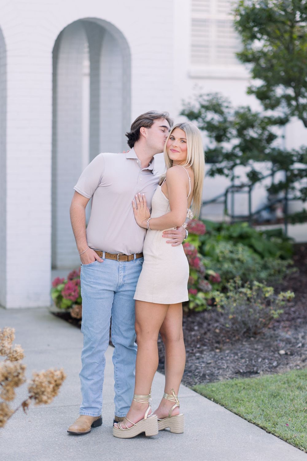 A romantic couple embraces on a garden pathway outside a white building during a photo session.