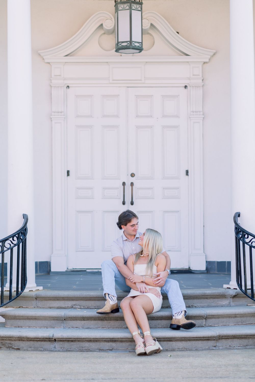 A couple sits together on stone steps in front of a grand white doorway with ornate architectural details and railings.