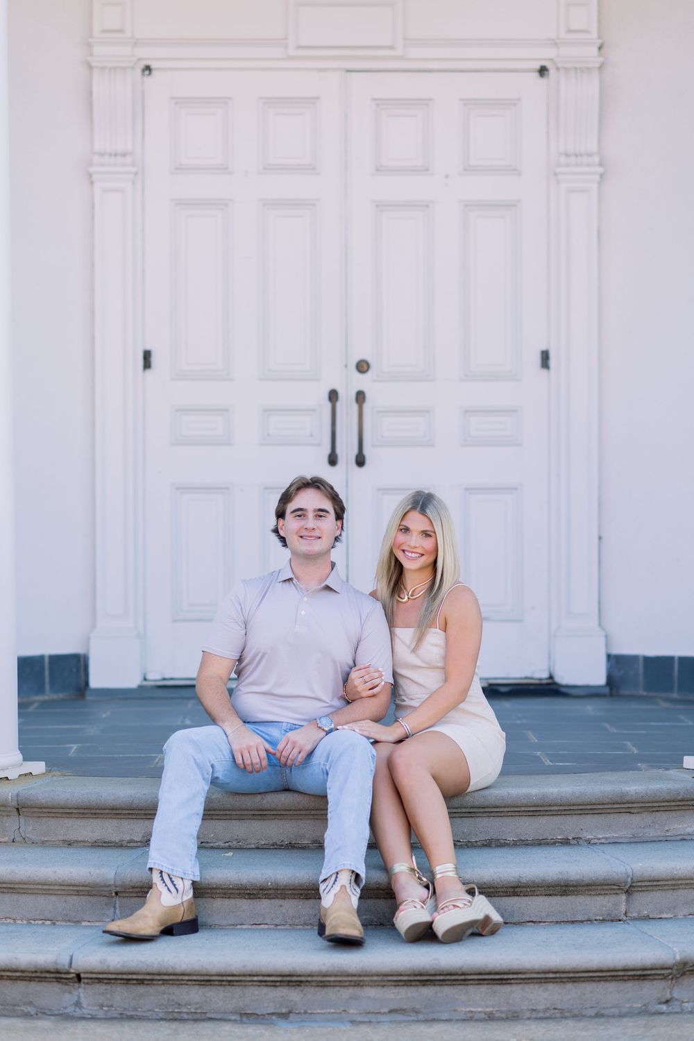A couple sits together on white stone steps in front of a classic white building entrance.