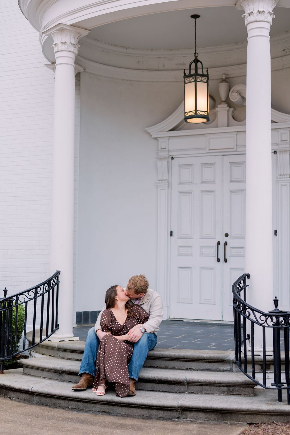 A couple sits together on white porch steps with columns and black railings in a romantic moment.