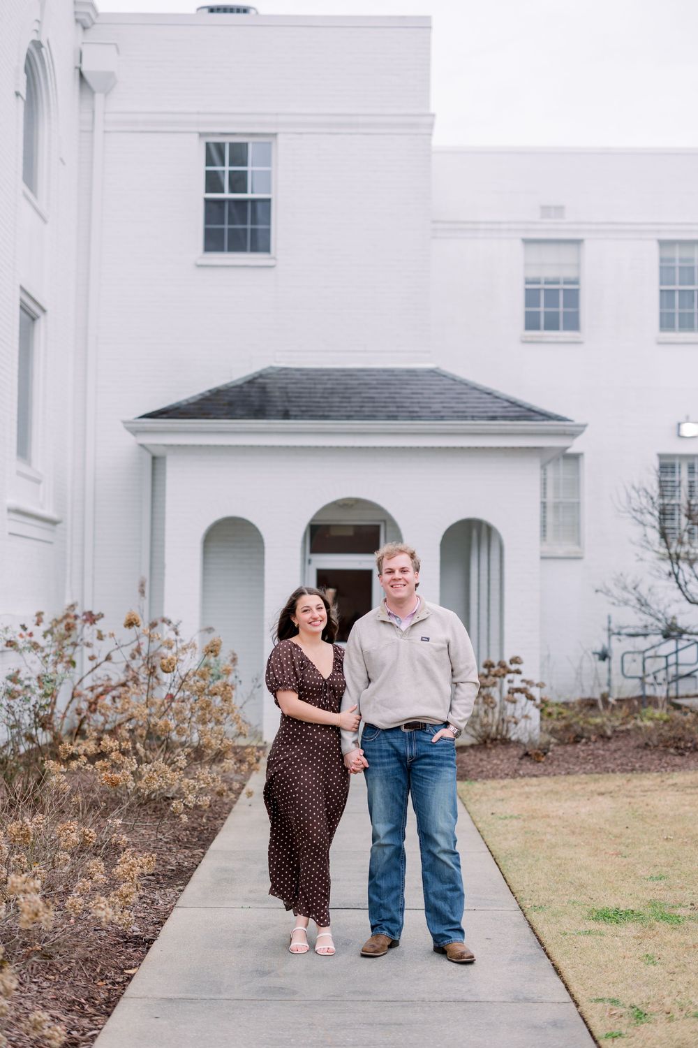 A couple stands together on a concrete walkway in front of a white building with arched entryway and classic architecture.
