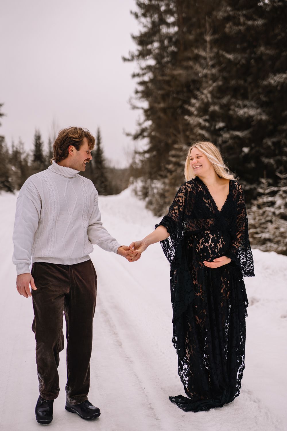 A couple holds hands while walking on a snowy path surrounded by evergreen trees, with her wearing a black lace dress.