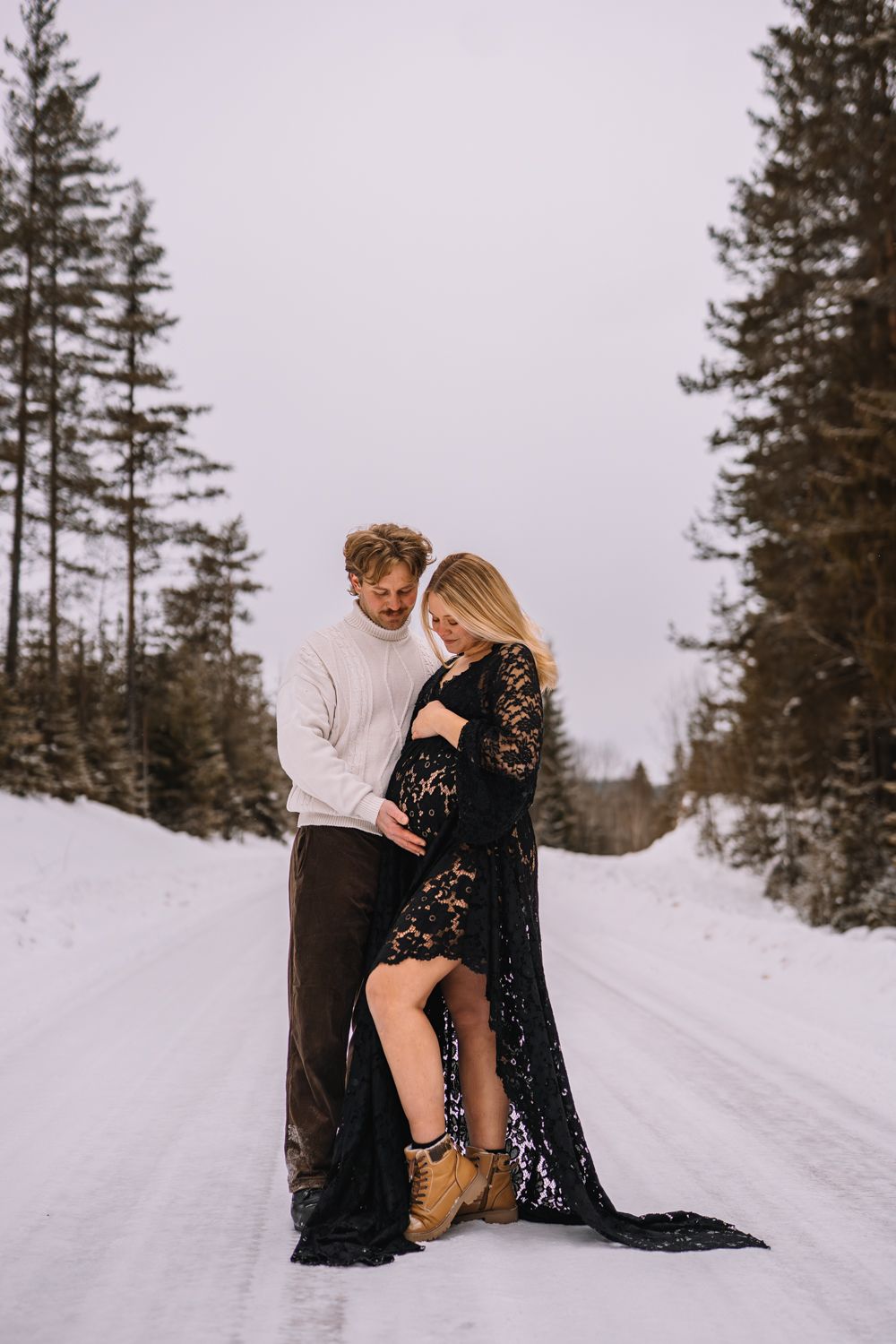 A couple embraces in a romantic winter setting with snow-covered evergreen trees in the background.