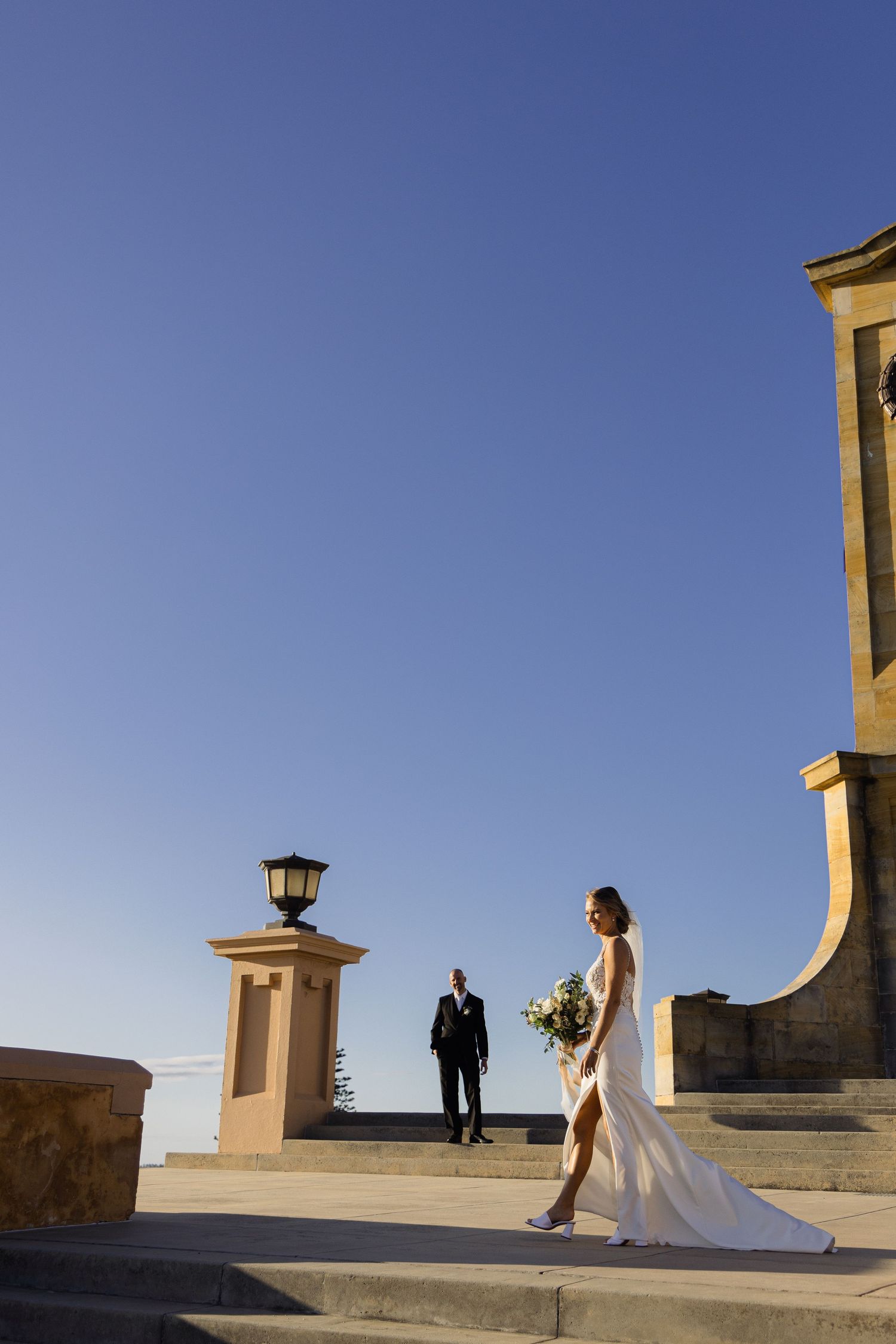 A bride and groom stand on stone steps against a bright blue sky at sunset, with architectural details in view.