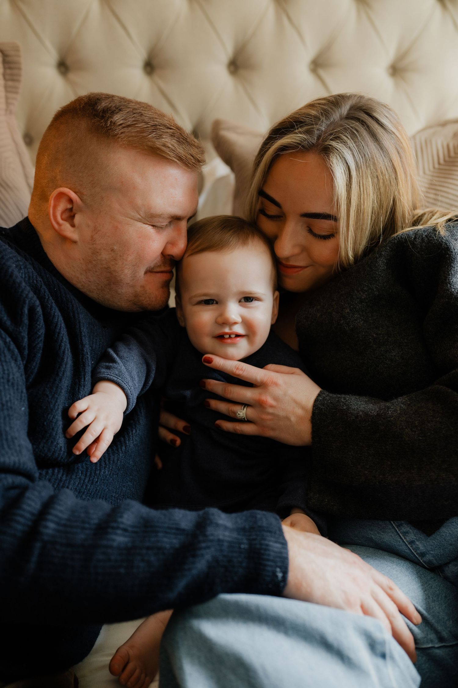 two parent kiss their baby boy head while sited on the bed