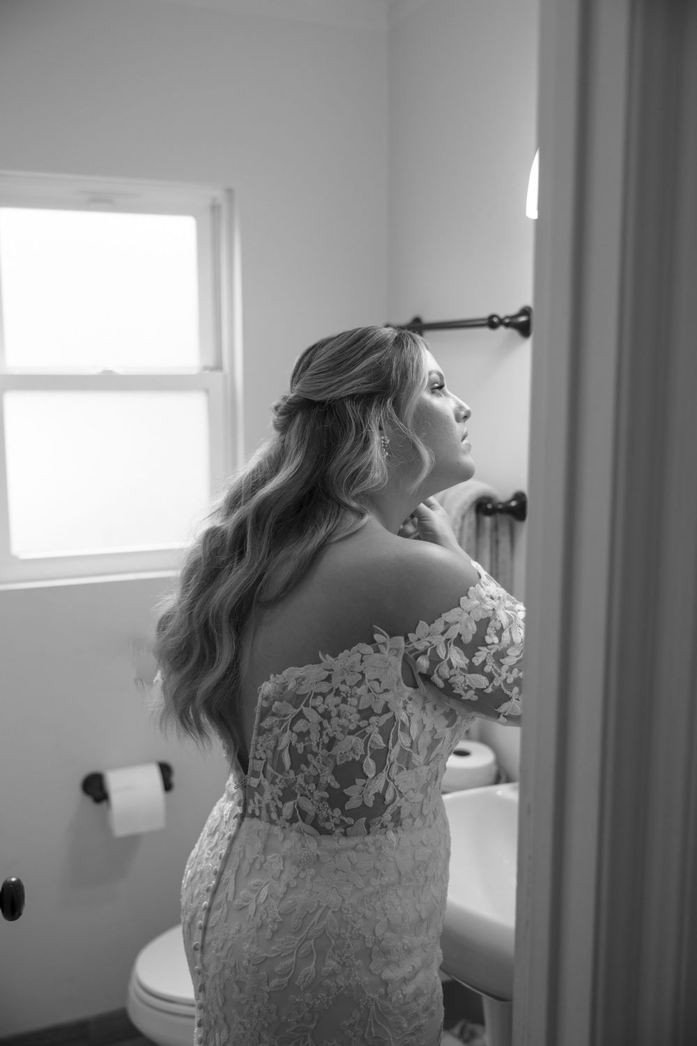 A bride in a white lace wedding dress stands by a window in black and white photography.