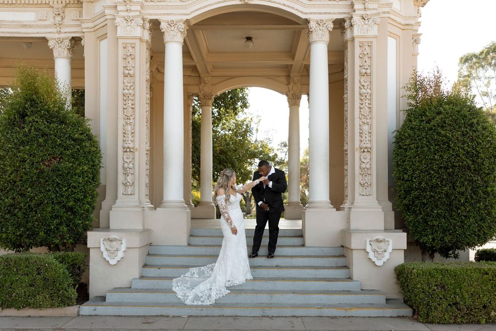A wedding couple stands on grand white stone steps with ornate columns and manicured hedges.