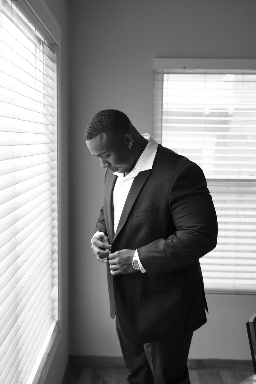 A groom adjusts his tie while getting ready near a bright window in black and white.