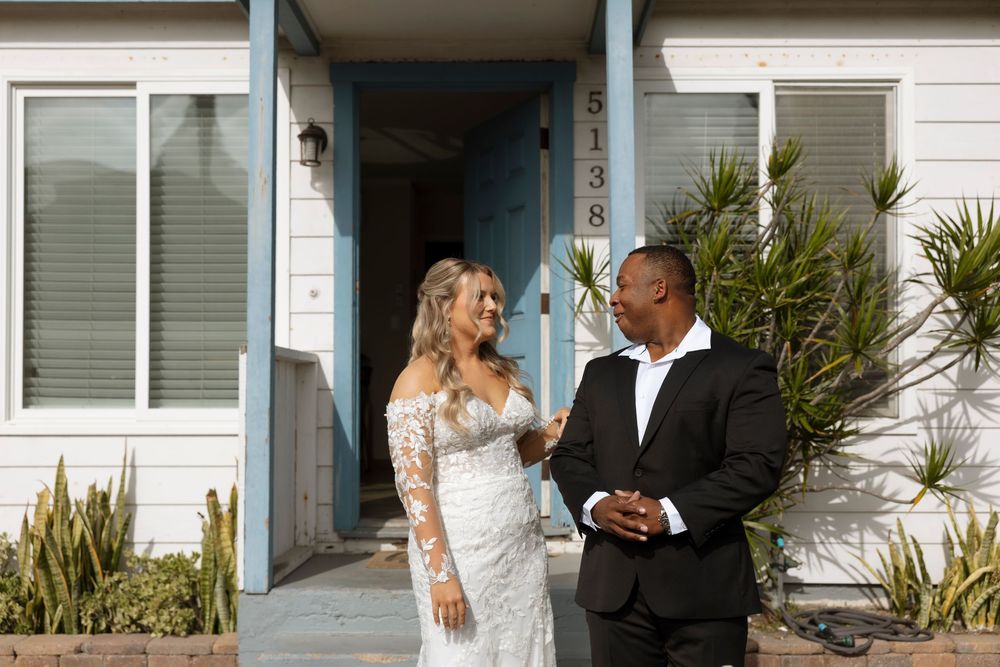 A couple in formal wedding attire shares a joyful moment together outside a white building with palm plants.