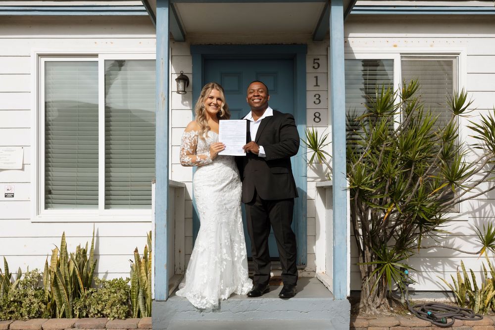 A wedding couple stands together in front of a white house entrance with palm plants visible.