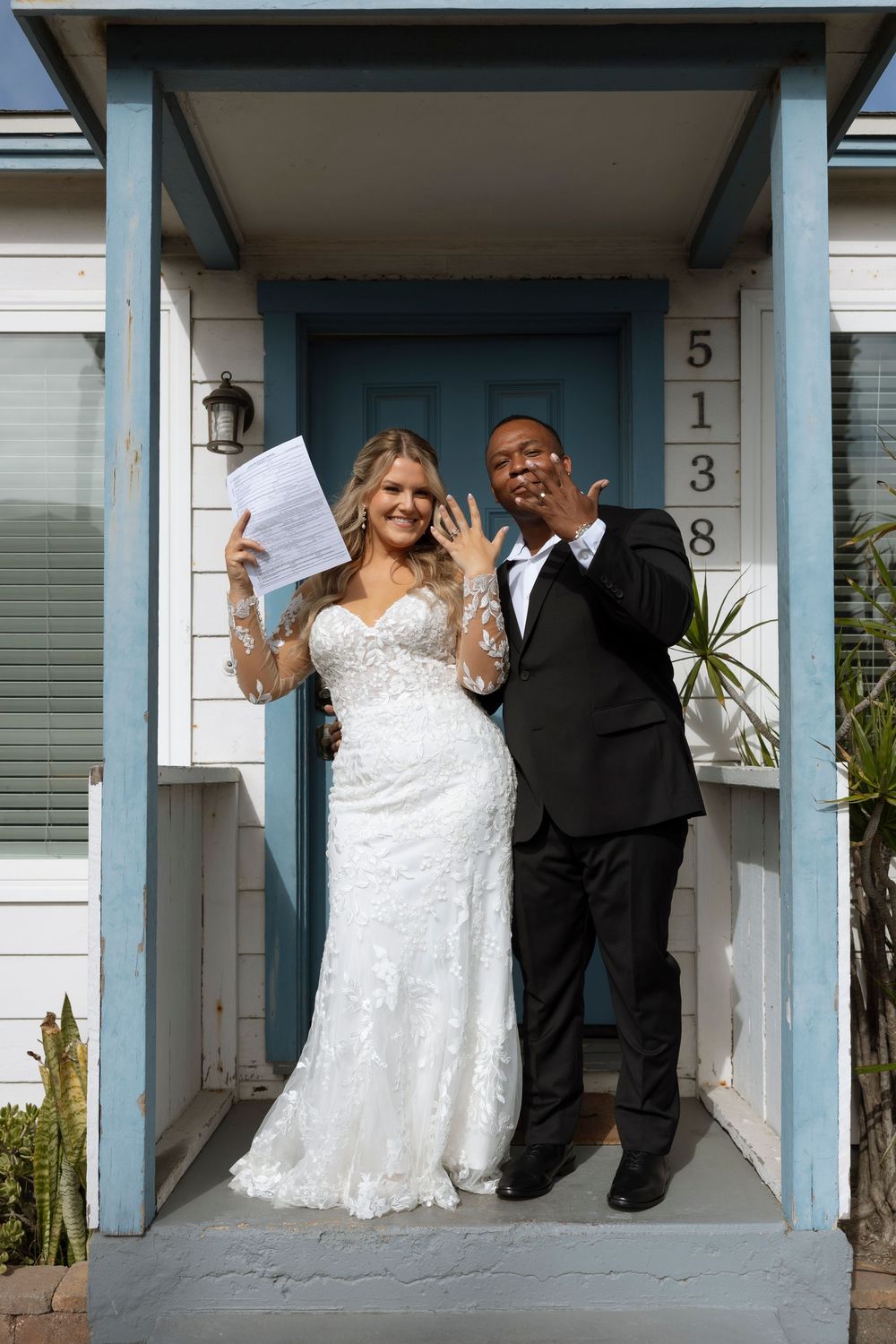 A wedding couple poses joyfully on a blue-trimmed front porch of a house with the number 5138.