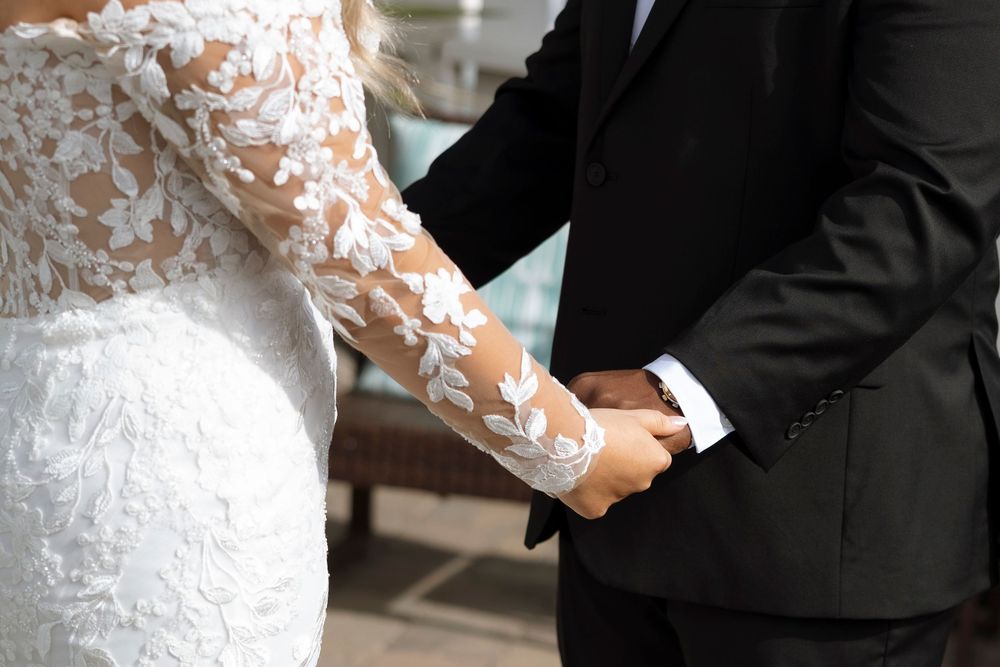 Close-up detail of a bride's delicate lace sleeve and hand holding a bouquet during an indoor wedding ceremony.