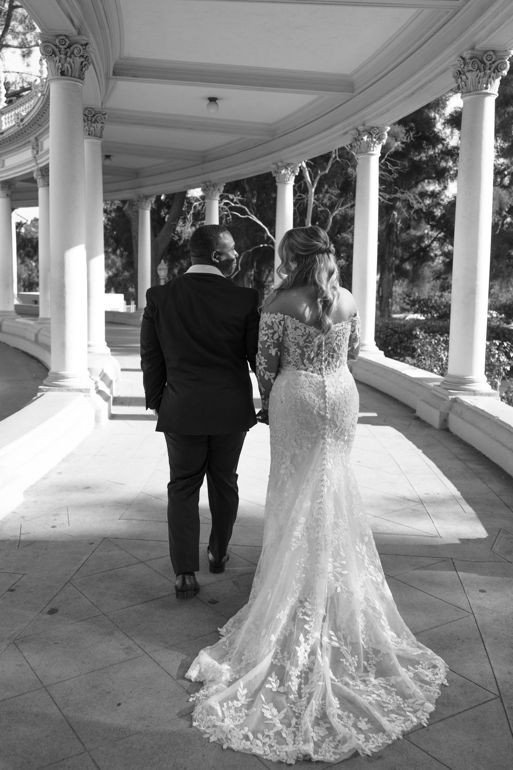 A couple in wedding attire walking along an elegant columned walkway in black and white.