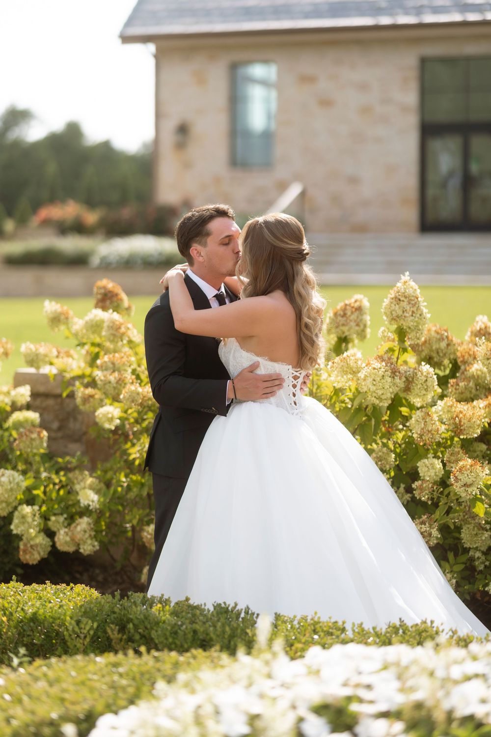 A romantic wedding couple embraces in a garden with blooming yellow hydrangeas and a stone building in the background.
