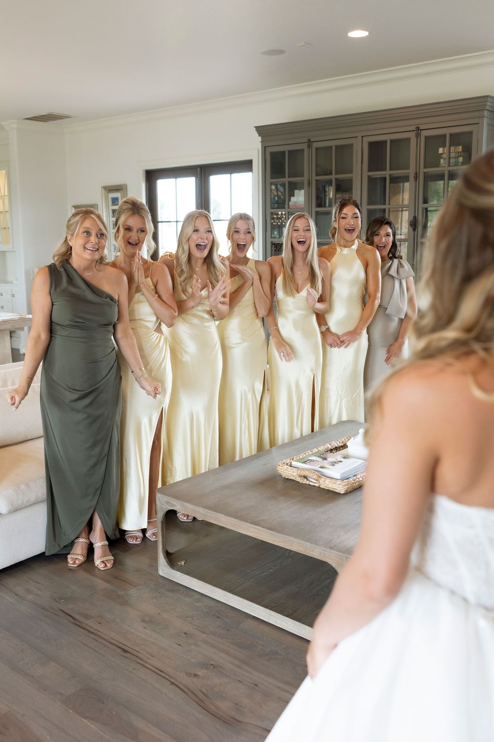 Bridesmaids in pale yellow dresses and groomsmen in gray suits gather for a wedding party photo in a bright hallway.