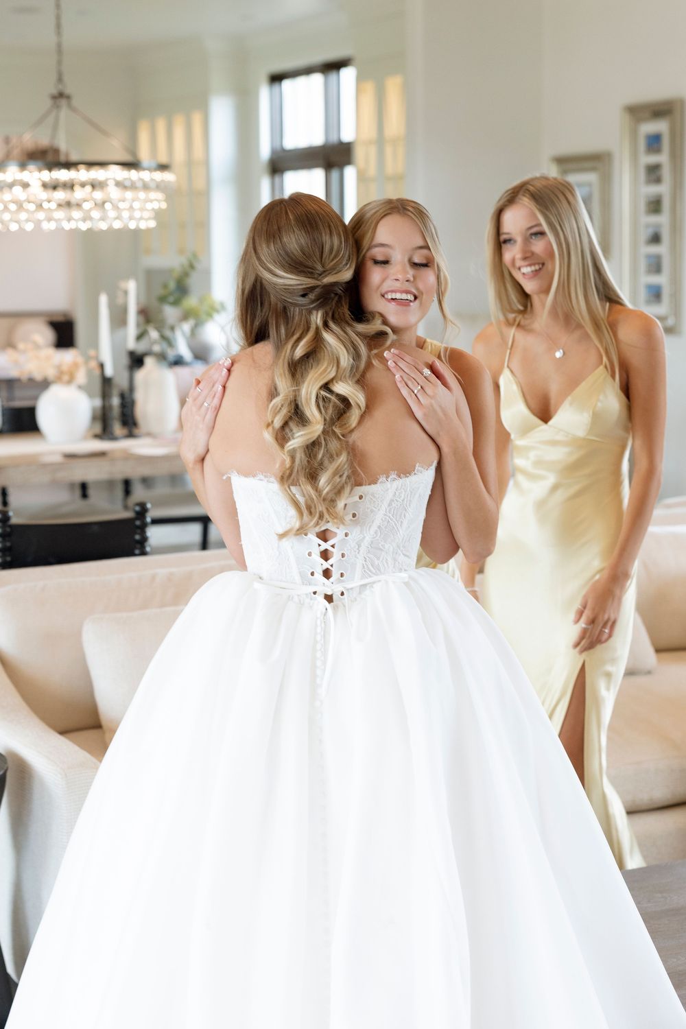 Two women in white and yellow dresses laugh together in an elegant room with chandelier lighting.