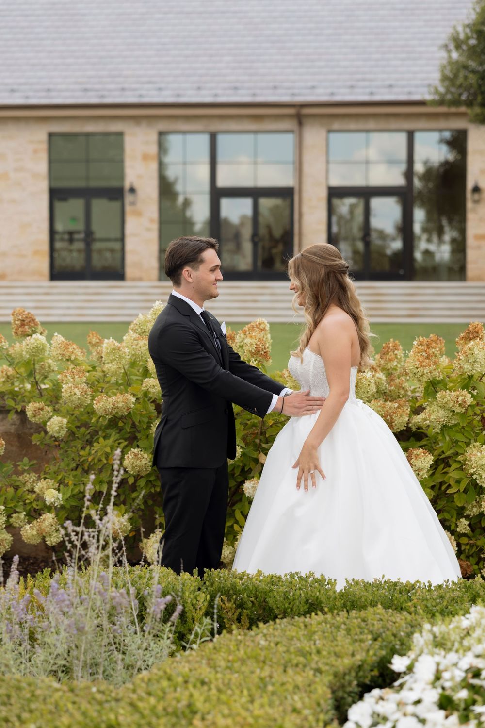 A wedding couple shares a playful moment among flowering bushes and gardens outside a stone building.